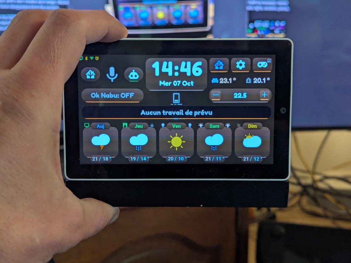 La M5Stack Tab5 de l'auteur tenue à la main sur son socle : l'heure, deux températures, la consigne de la clim, le bandeau des horaires de travail et les prévisions à 5 jours avec orage, pluie et soleil